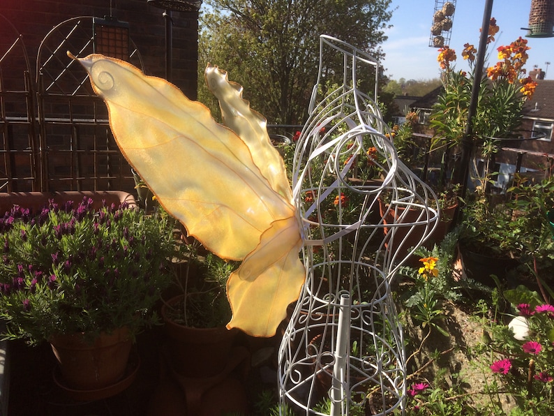 May include: A pair of large, iridescent, yellow fairy wings on a white wire dress form. The wings are made of a translucent material and have a delicate, ethereal look.