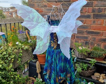 May include: White fairy wings with a green and blue pattern on a dress form. The wings are made of sheer fabric and have a delicate, ethereal look.
