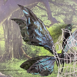 May include: A pair of large, iridescent black and green wings on a white wire dress form. The wings are made of fabric and have a textured, almost scaly appearance.
