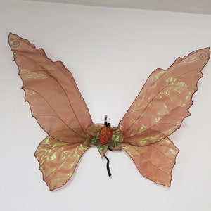 May include: Brown and gold butterfly wings with a shimmering, iridescent effect. The wings are made of a sheer fabric with a detailed vein pattern. The center features a decorative element and black straps.