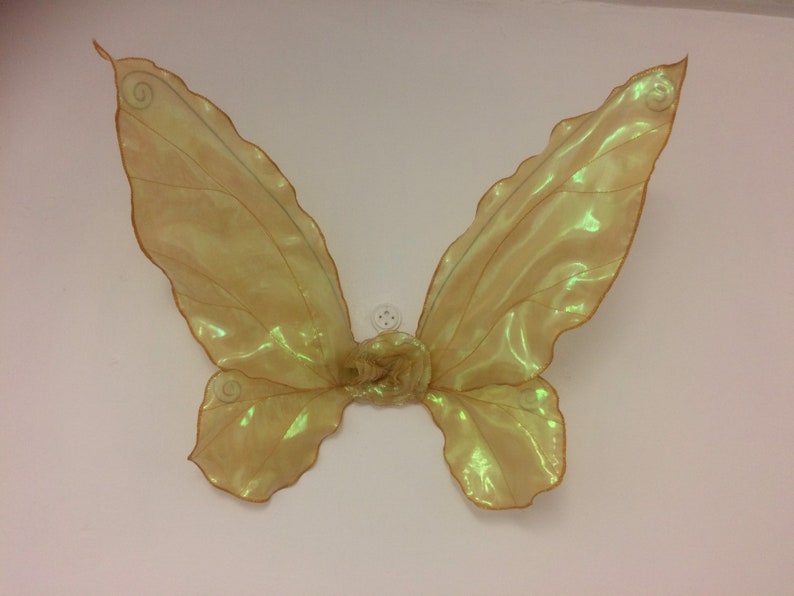May include: A pair of iridescent gold fairy wings with a delicate, sheer fabric. The wings are attached at the center with a small, white button.