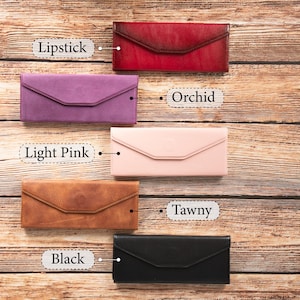 Puede incluir: Cinco carteras de cuero de diferentes colores dispuestas sobre una superficie de madera. Las carteras están etiquetadas como "Lipstick", "Orchid", "Light Pink", "Tawny" y "Black".