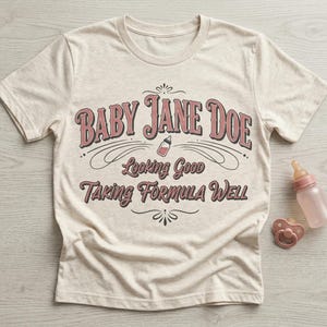 The pitt Baby Jane Doe funny slogan t shirt with baby bottle graphic, cream graphic tee flat lay with baby bottle and pacifier props on wooden background, styled product image highlighting vintage typography humor design inspired by the pitt aesthetic and modern casual fashion trend