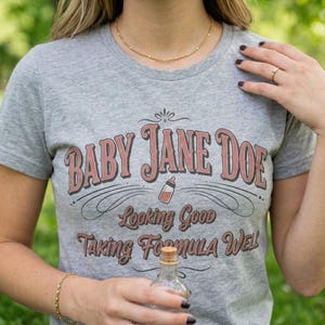 The Pitt Baby Jane Doe funny slogan t shirt with baby bottle graphic, woman wearing gray graphic tee outdoors holding small bottle, casual lifestyle image with natural greenery background, vintage typography humor design inspired by the pitt aesthetic and modern everyday fashion trend