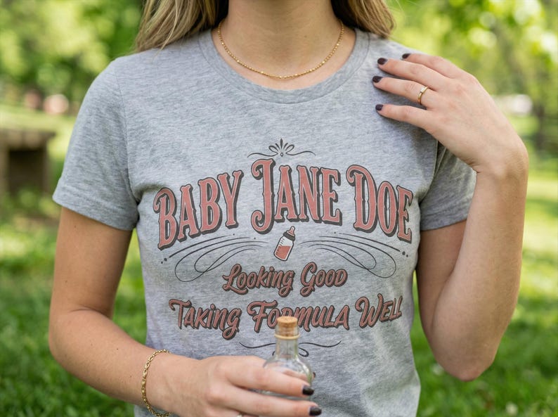 The Pitt Baby Jane Doe funny slogan t shirt with baby bottle graphic, woman wearing gray graphic tee outdoors holding small bottle, casual lifestyle image with natural greenery background, vintage typography humor design inspired by the pitt aesthetic and modern everyday fashion trend
