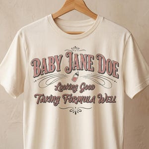 The Pitt Baby Jane Doe funny slogan t shirt with baby bottle graphic, cream graphic tee hanging on wooden hanger against neutral background, clean minimal product image showcasing vintage typography humor design inspired by the pitt aesthetic and modern casual fashion trend