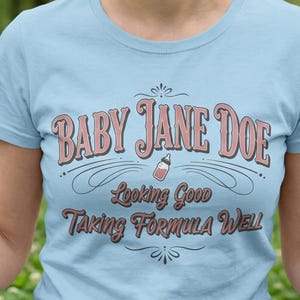 The Pitt Baby Jane Doe funny slogan t shirt with baby bottle graphic, woman wearing blue graphic tee outdoors in park setting, casual lifestyle image with soft natural greenery background, vintage typography humor design inspired by the pitt aesthetic and modern everyday fashion trend
