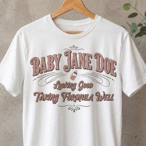 The Pitt Baby Jane Doe funny slogan t shirt with baby bottle graphic, white graphic tee hanging on wooden hanger with minimal decor background, clean product display image highlighting vintage typography humor design inspired by the pitt aesthetic and modern casual fashion trend