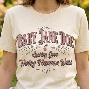 The Pitt Baby Jane Doe funny slogan t shirt with baby bottle graphic, woman wearing cream graphic tee outdoors in sunny park setting, casual lifestyle image with soft natural background, vintage typography humor design inspired by the pitt aesthetic and modern everyday fashion trend
