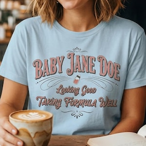 The Pitt Baby Jane Doe funny slogan t shirt with baby bottle graphic, woman wearing soft blue graphic tee in cozy coffee shop holding latte, aesthetic lifestyle image with vintage typography humor design inspired by the pitt trend and casual everyday fashion style