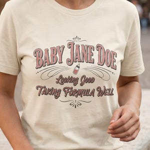 The Pitt Baby Jane Doe funny slogan t shirt with baby bottle graphic, woman wearing soft cream graphic tee walking outdoors, casual lifestyle street photo with vintage typography humor design inspired by the pitt aesthetic and modern everyday fashion style