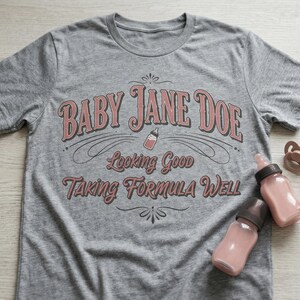 The Pitt Baby Jane Doe funny slogan t shirt with baby bottle graphic, gray graphic tee flat lay with baby bottles and pacifier props on wooden background, styled product image highlighting vintage typography humor design inspired by the pitt aesthetic and modern casual fashion trend