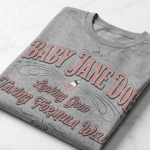 The Pitt Baby Jane Doe funny slogan t shirt with baby bottle graphic, folded gray graphic tee flat lay on marble background, clean product mockup highlighting vintage typography humor design inspired by the pitt aesthetic and modern casual fashion trend