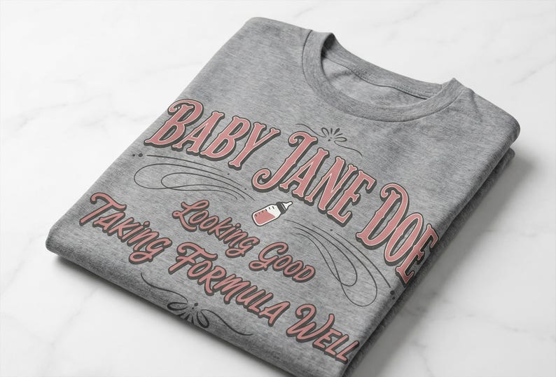 The Pitt Baby Jane Doe funny slogan t shirt with baby bottle graphic, folded gray graphic tee flat lay on marble background, clean product mockup highlighting vintage typography humor design inspired by the pitt aesthetic and modern casual fashion trend