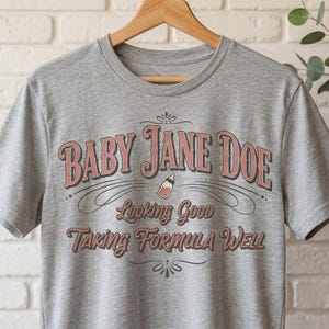 The pitt Baby Jane Doe funny slogan t shirt with baby bottle graphic on gray tee, hanging on wooden hanger against white brick wall, trendy vintage typography design, humorous mom shirt gift, casual unisex graphic tee for everyday wear, soft neutral aesthetic