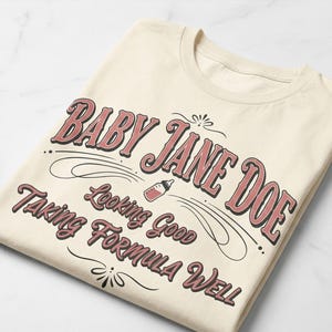 The Pitt Baby Jane Doe funny slogan t shirt with baby bottle graphic, folded cream graphic tee flat lay on clean background, product display image highlighting vintage typography humor design inspired by the pitt aesthetic and trendy casual fashion style