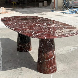 May include: A large oval dining table crafted from deep red marble with prominent white veining. The table features a smooth, polished surface and is supported by two substantial, matching conical marble pedestals in the same red stone.