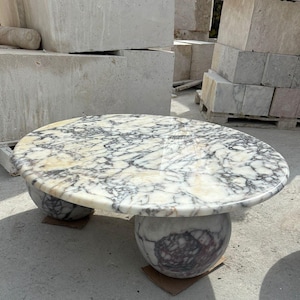 Op de afbeelding: Een witte marmeren salontafel met een rond blad en drie poten. De tafel heeft een uniek ontwerp met een draaiend patroon van wit, grijs en zwart marmer.