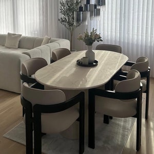 May include: A modern dining room featuring a light-colored oval table with six chairs. The chairs have black frames and beige seats. A black chandelier hangs above the table. A beige sectional sofa and two coffee tables are in the background.