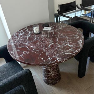 May include: A round marble table with a red and white veined pattern, supported by a pedestal base. Two black chairs with curved arms and a textured fabric surround the table. A candle and a small box are on the table.