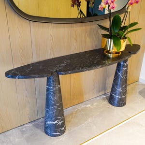 May include: A modern, black marble console table with two cone-shaped legs. The table has a unique, asymmetrical design and is topped with a gold planter holding a flowering plant. A brown leather ottoman sits nearby.