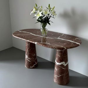 May include: An oval marble table in a deep burgundy colour with white veining, supported by two cylindrical legs. A clear glass vase holds white lilies. The table's dimensions are approximately 150 cm in length and 60 cm in width, a stylish piece.