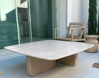 May include: A square, light-colored stone coffee table with rounded edges and two rectangular supports. The table is outdoors, with wooden chairs and a small ceramic side table visible in the background.
