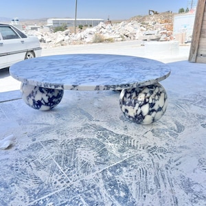 May include: A round marble coffee table with a white and gray patterned top. The table is supported by two spherical bases with a similar black and white pattern. The table is outdoors on a concrete surface.