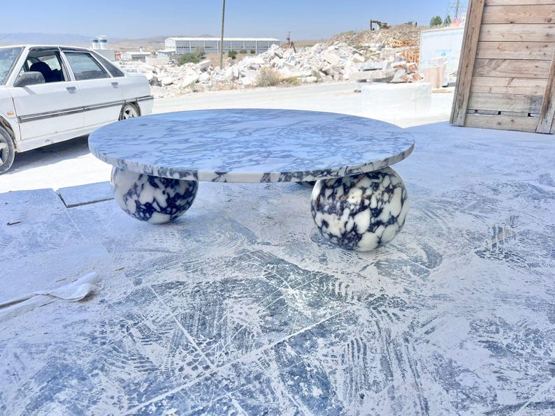 May include: A round marble coffee table with a white and gray patterned top. The table is supported by two spherical bases with a similar black and white pattern. The table is outdoors on a concrete surface.