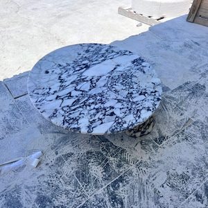May include: A round marble table with a white and dark blue veined pattern. The table has a pedestal base and is set on a concrete surface. The table's diameter is approximately 36 inches.