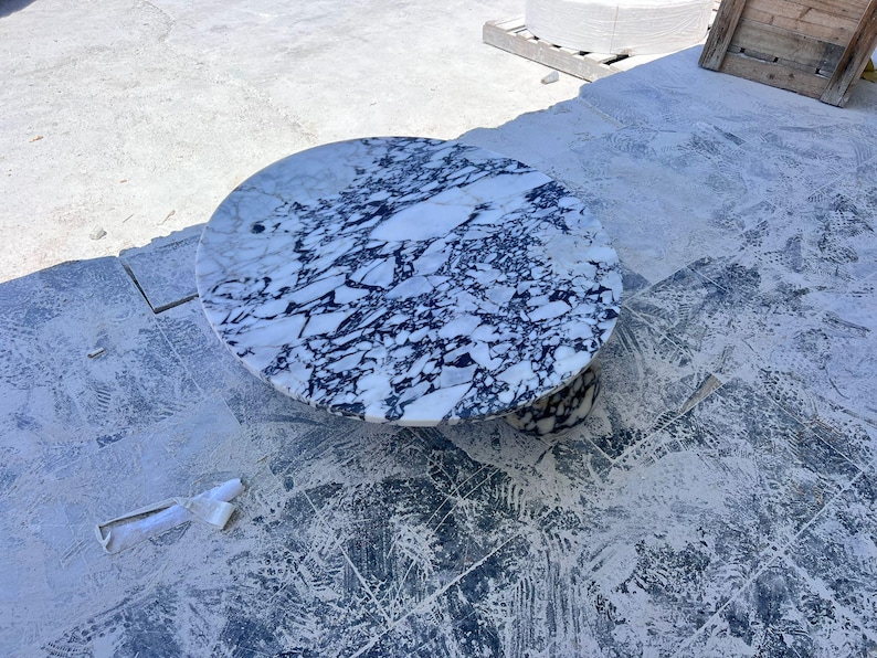 May include: A round marble table with a white and dark blue veined pattern. The table has a pedestal base and is set on a concrete surface. The table's diameter is approximately 36 inches.