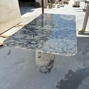 Puede incluir: Una mesa rectangular de mármol con borde redondeado. La encimera presenta un patrón veteado blanco y gris. La mesa está sostenida por una base cilíndrica de mármol a juego. La mesa mide aproximadamente 183 cm de largo y 91 cm de ancho.