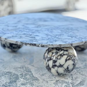 May include: A round marble table with a white and gray pattern. The tabletop rests on three spherical legs, also made of marble. The table is a decorative piece, suitable for various interior settings.
