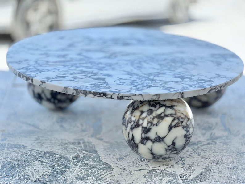 May include: A round marble table with a white and gray pattern. The tabletop rests on three spherical legs, also made of marble. The table is a decorative piece, suitable for various interior settings.