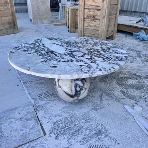 May include: A round marble table with a white and black patterned top. The table is supported by a spherical base with similar veining. The table is a statement piece for any living space.