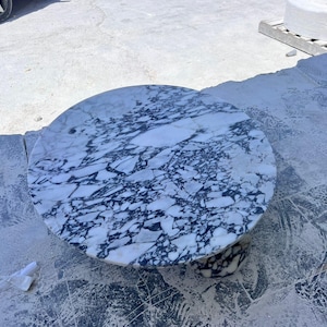May include: A round marble table with a white and dark blue veined pattern. The table has a circular top and a pedestal base. The table is likely for indoor or outdoor use.