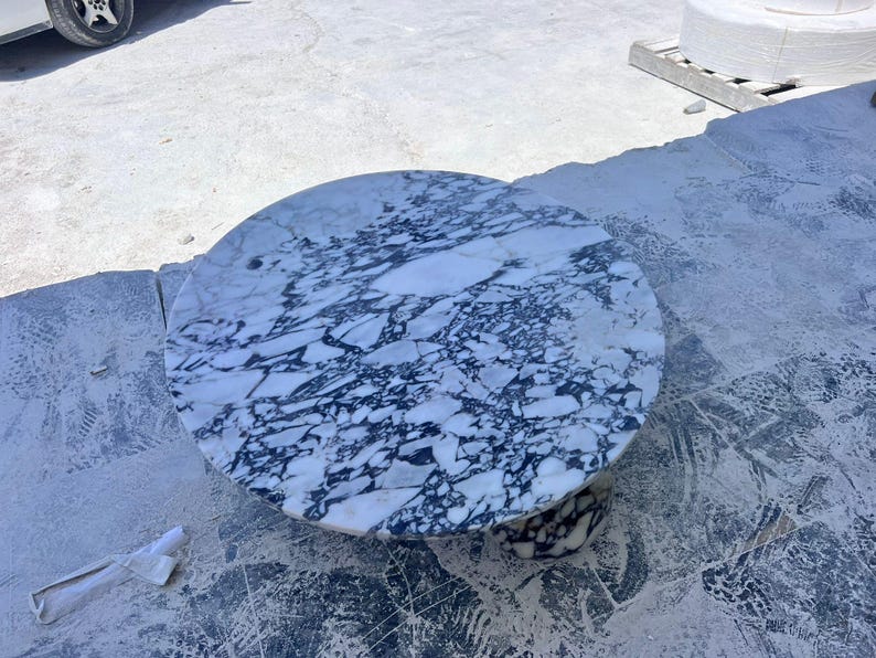 May include: A round marble table with a white and dark blue veined pattern. The table has a circular top and a pedestal base. The table is likely for indoor or outdoor use.