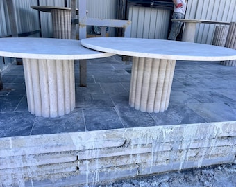 May include: Two oval-shaped, white marble tables with fluted bases. The tables are on a gray tiled floor with a white stone wall behind them.