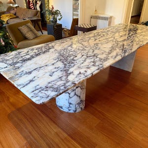 Pode incluir: Uma mesa de jantar retangular de mármore com um padrão branco e roxo. A mesa é suportada por uma base cilíndrica. A mesa está sobre um piso de madeira.