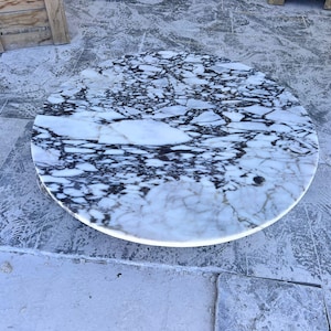 May include: A round marble table with a white and black patterned surface. The table has a polished finish and is set against a gray background. The table's design is a striking contrast of light and dark tones.