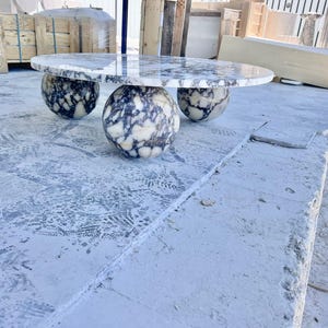 May include: A modern oval marble table with three spherical legs. The tabletop features a white and gray marbled pattern. The legs have a similar pattern. The table is set on a concrete surface.