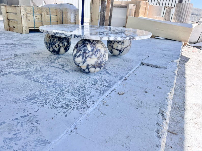 May include: A modern oval marble table with three spherical legs. The tabletop features a white and gray marbled pattern. The legs have a similar pattern. The table is set on a concrete surface.