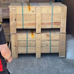 May include: Two large wooden crates stacked inside a delivery vehicle. The crates are secured with green straps and have the DHL logo visible. Other wooden crates are stacked in the background.