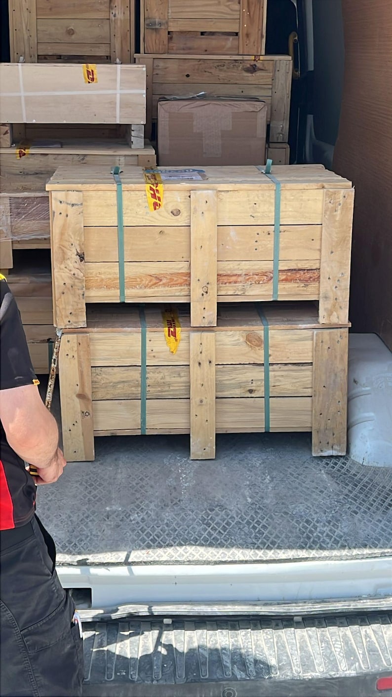 May include: Two large wooden crates stacked inside a delivery vehicle. The crates are secured with green straps and have the DHL logo visible. Other wooden crates are stacked in the background.