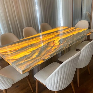 May include: A rectangular dining table with a glowing, multi-colored onyx top. The table is surrounded by eight white upholstered chairs with wooden legs. The table's surface has shades of yellow, orange, and gray.