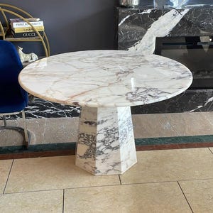 Op de afbeelding: Een ronde witte marmeren eettafel met een geometrische basis. Het tafelblad heeft een draaiend patroon van grijze en bruine aderen. De tafel staat op een beige betegelde vloer.