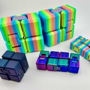 Può includere: Diversi giocattoli fidget colorati sono esposti su una superficie bianca. I giocattoli includono cubi a strisce arcobaleno e blocchi rettangolari in varie combinazioni di colori, tra cui blu, verde, rosa e viola. Questi giocattoli sono progettati per alleviare lo stress e il gioco sensoriale.