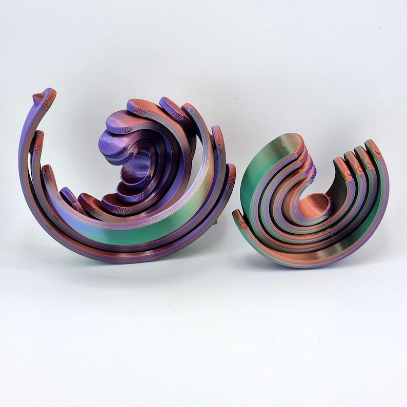 May include: Two abstract sculptures in a wave-like design. The larger sculpture is a mix of purple, green, and orange hues, while the smaller one features green and orange tones. Both are set against a white background.