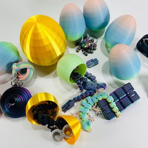 Könnte beinhalten: Eine Sammlung von 3D-gedruckten Spielzeugen und Dekorationsartikeln in verschiedenen Formen und Farben. Enthält eiförmige Objekte mit Farbverläufen, eine gelbe Kugel und verschiedene Zappelspielzeuge. Die Artikel sind auf einer weißen Oberfläche angeordnet.
