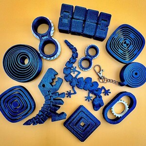 Könnte beinhalten: Eine Sammlung blauer Fidget-Spielzeuge, darunter ein segmentierter Block, ein Dinosaurier, eine Eidechse und verschiedene kreisförmige und geometrische Formen. Einige Spielzeuge haben weiße Akzente. Ein Schlüsselanhänger ist ebenfalls enthalten.